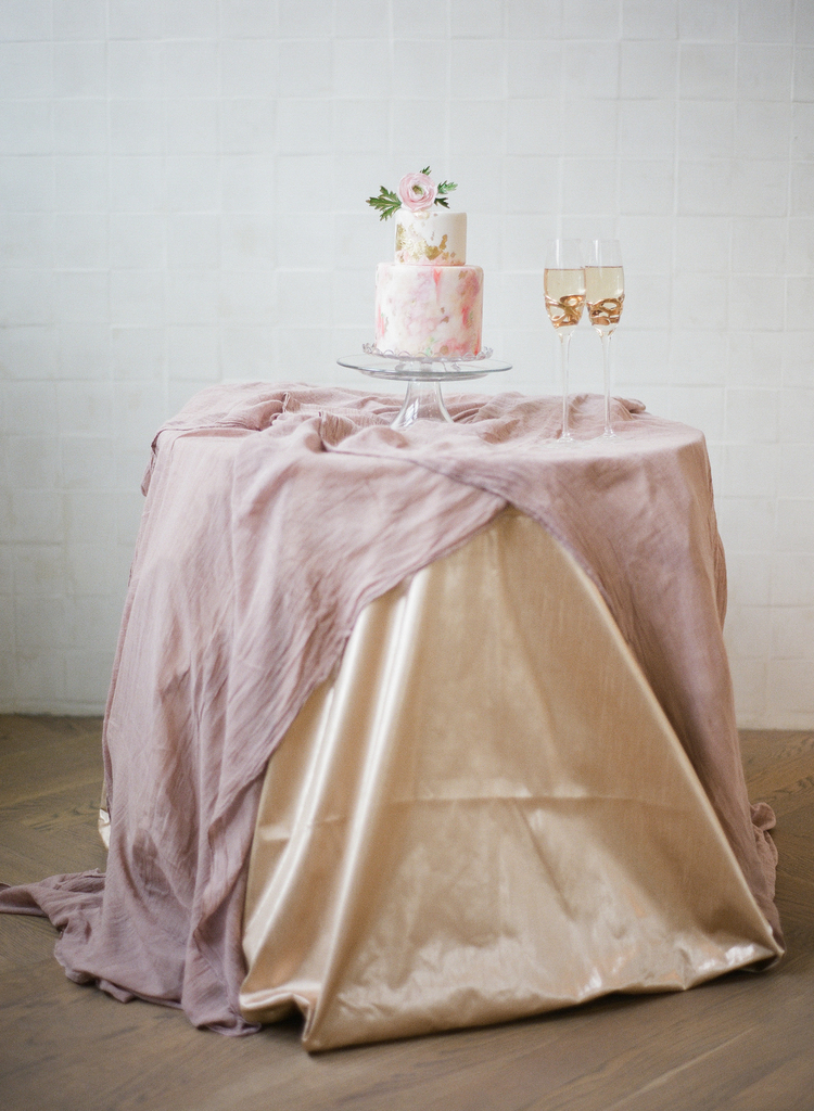 two tier Pink and gold wedding cake with a pink flower on top next to two glasses of champagne on top of a blush linen