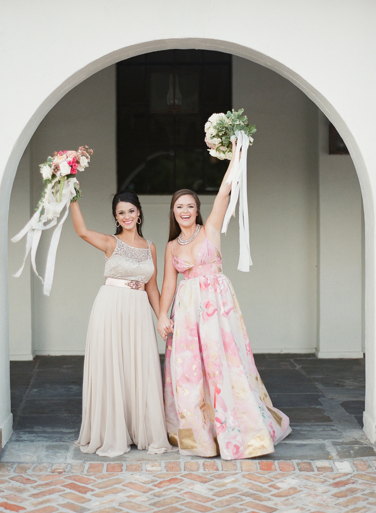 Brides holding up their bouquets smiling outside under an archway 