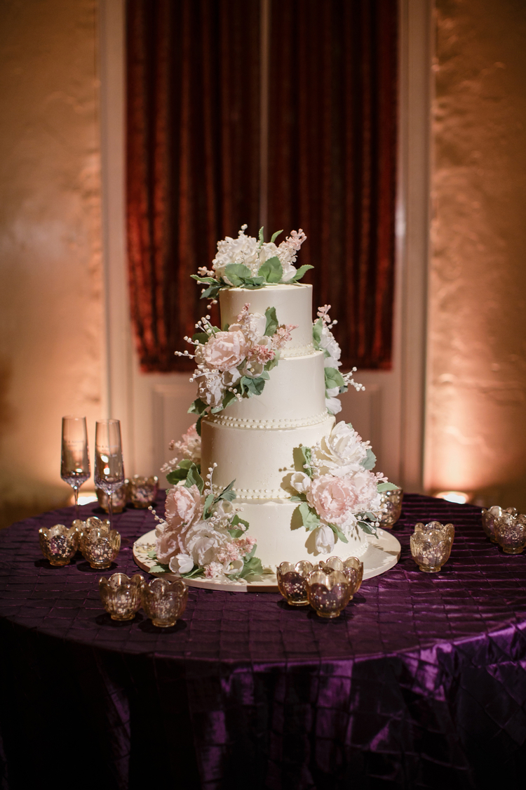 Four tier white wedding cake with pink and white flowers placed on each tier sitting on a plum purple table linen