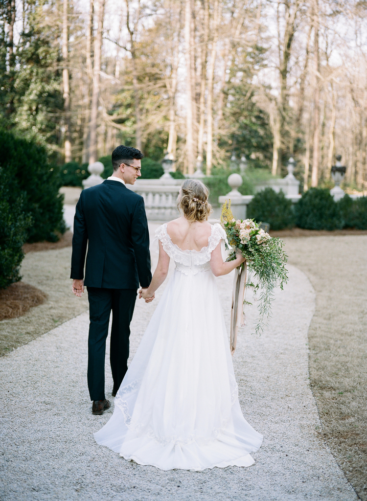 Bride and groom walking down a gravel path holding out her lush greenery bouquet with nude sashes hanging 