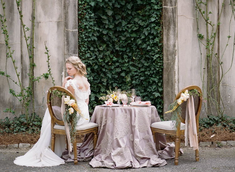 Sweetheart table with a blush linen and chairs with greenery and floral attached  in front of an ivy wall with the bride in a chair 