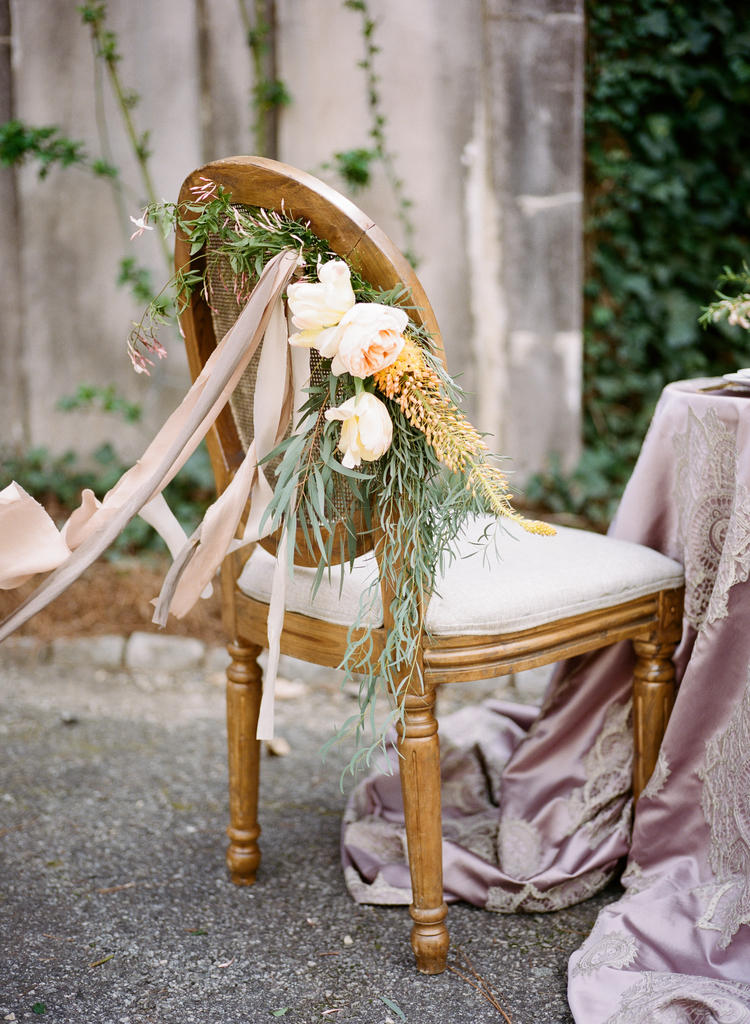 Close up of the chairs with ribbons, greenery, peonies and roses ties to the back next to a table with bush linens