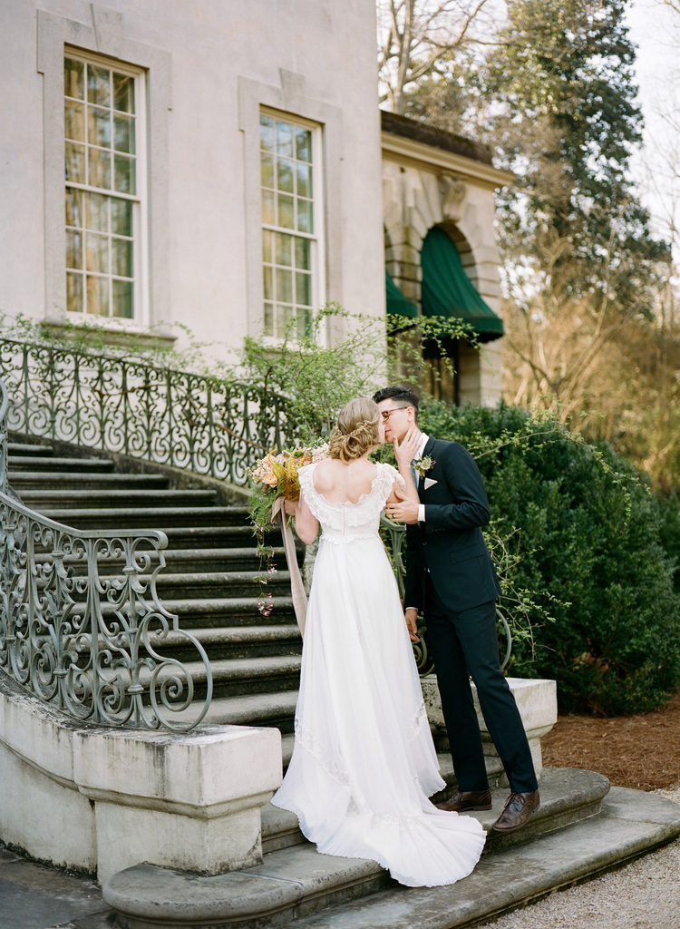 bride and groom kiss at the bottom of a stair case with green bushes in the background