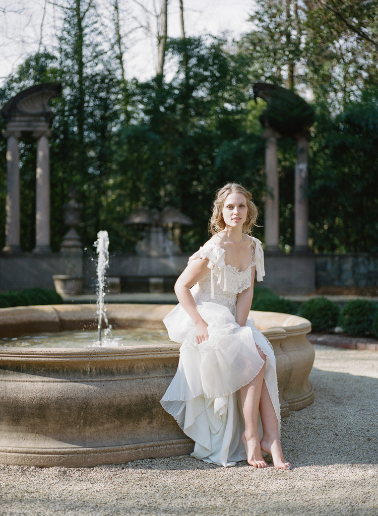 Bride in a lace wedding gown sits on a round fountain barefoot in the gravel