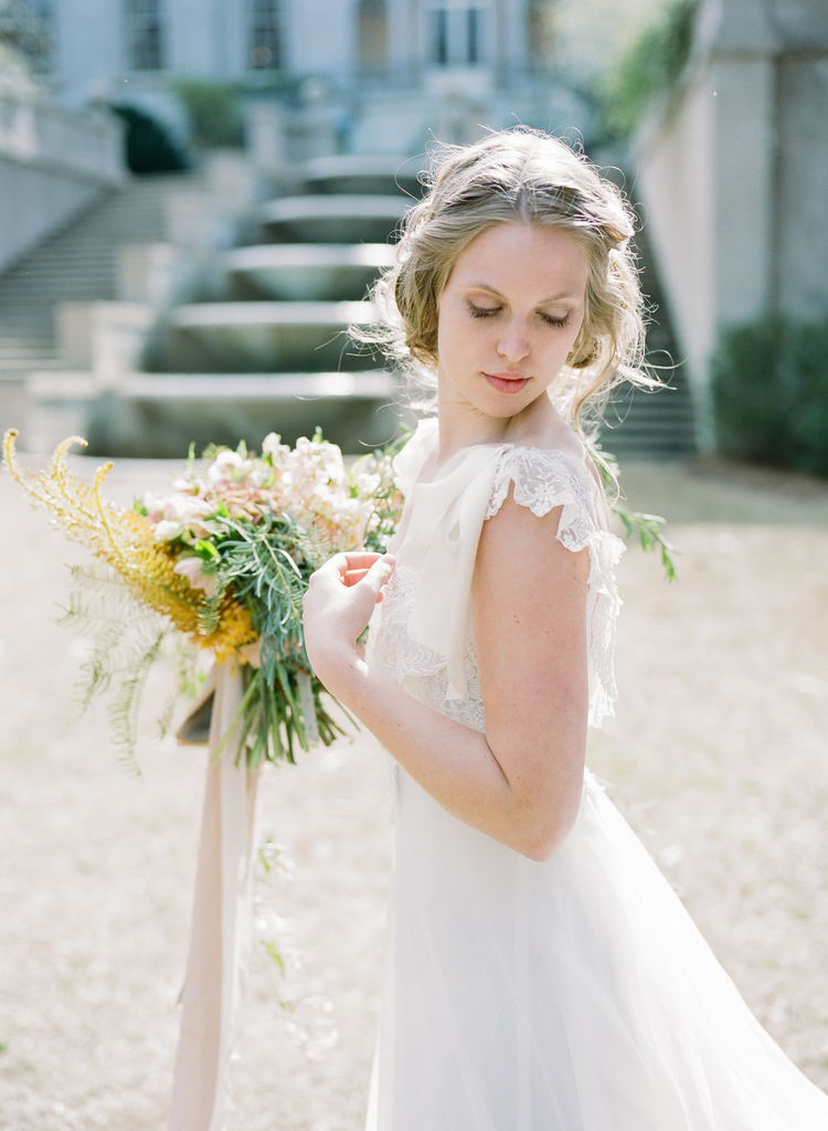 a bride in her wedding gown holding her wedding bouquet of yellow, pink and green floral looks over her shoulder