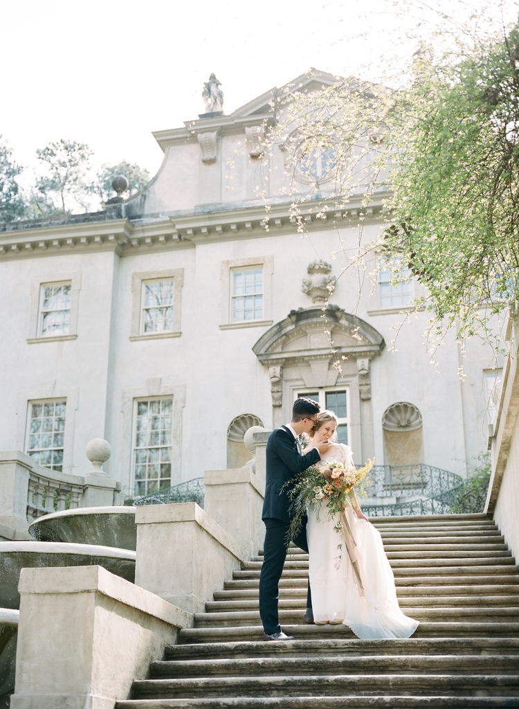 Bride and groom standing on steps leading to a mansion laughing in their wedding attire and lush floral bouquet 