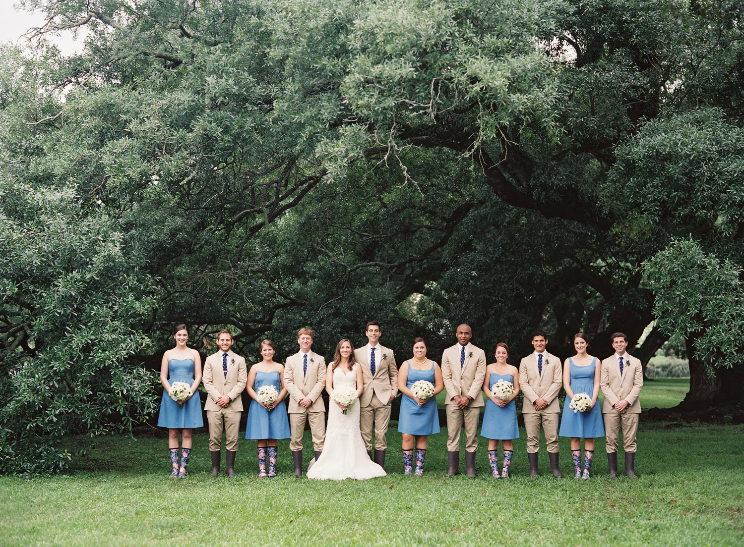 Bride, groom, bridesmaids, groomsmen all lined up in their rain-boots on a rainy wedding day in front of oak trees 