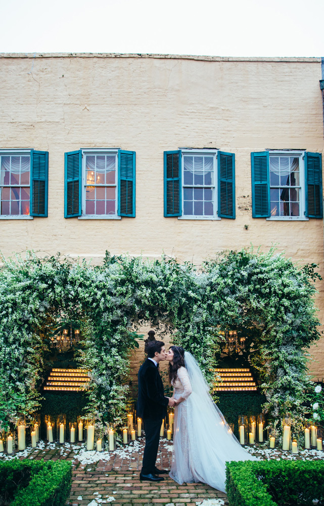 Bride and groom hold hands and kiss in front of greenery draping behind them and candles flickering with white rose petals on the ground 