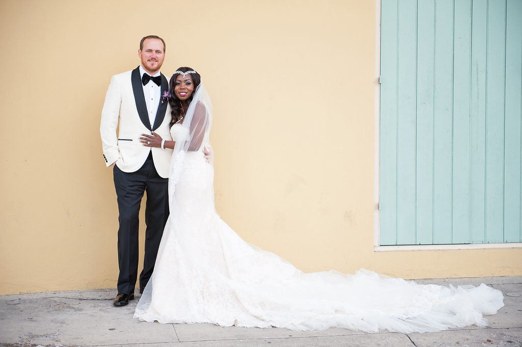 bride and groom holding each other standing in front of a yellow wall with green shutters smiling at the camera